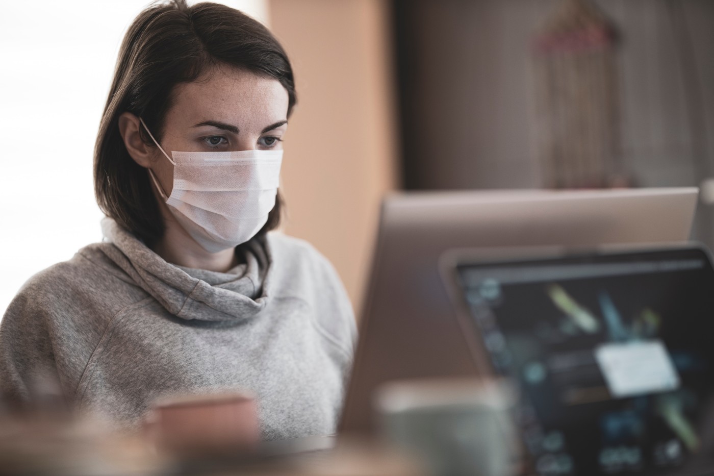 A woman in a grey sweater wearing a face mask while working at a computer.